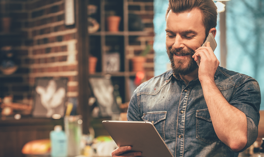 our appointment is on Monday! Cheerful young bearded man talking on mobile phone and looking at digital tablet while standing at barbershop