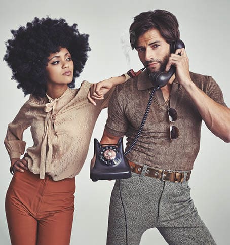 A beautiful young hippie looking over at a handsome man using a retro telephone.