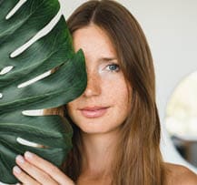 Young beautiful woman covering her face with green leaf at home. Beauty and skin care concept. Rejuvenating anti-age effect for hair and body.