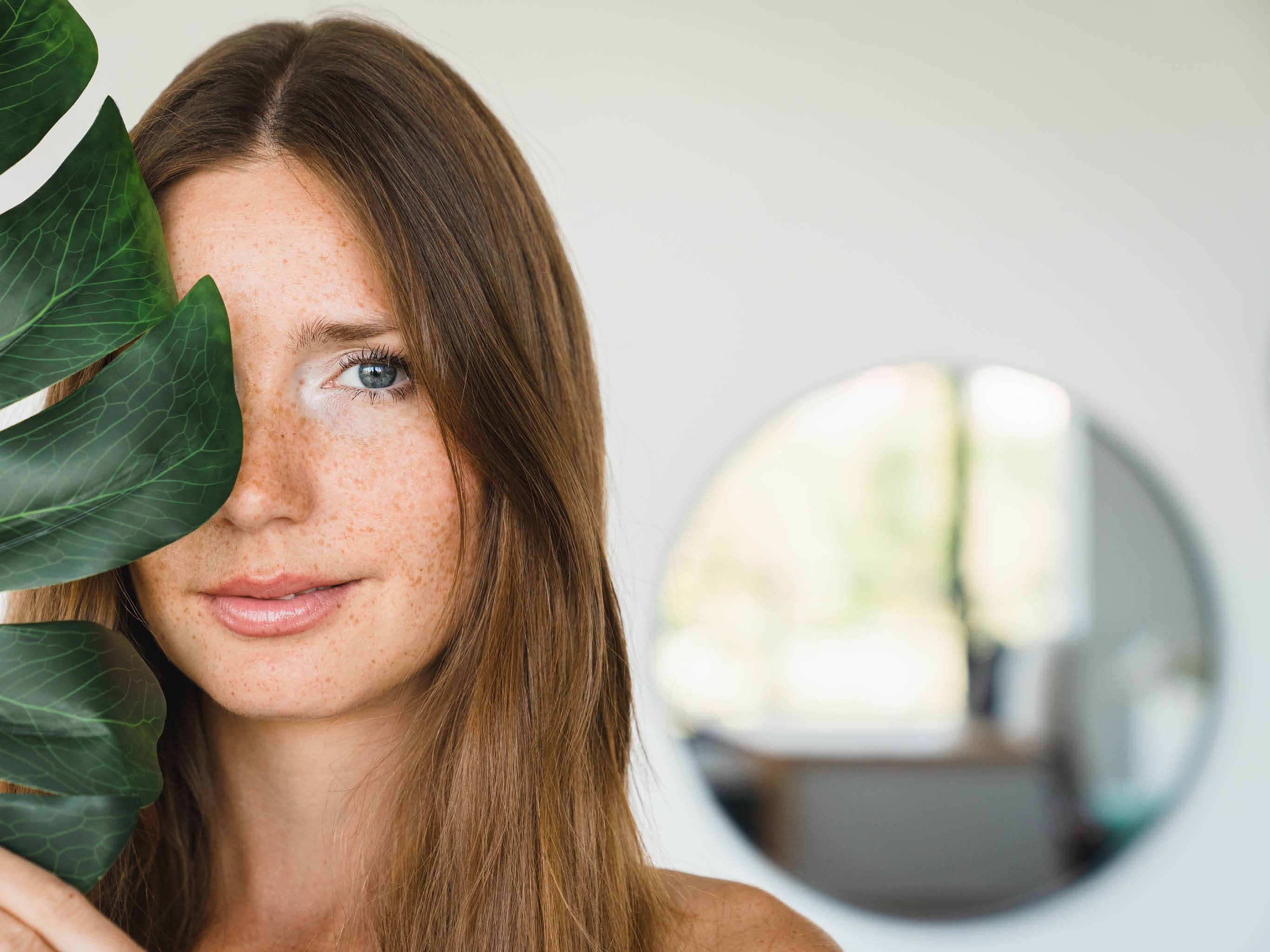 Young beautiful woman covering her face with green leaf at home. Beauty and skin care concept. Rejuvenating anti-age effect for hair and body.