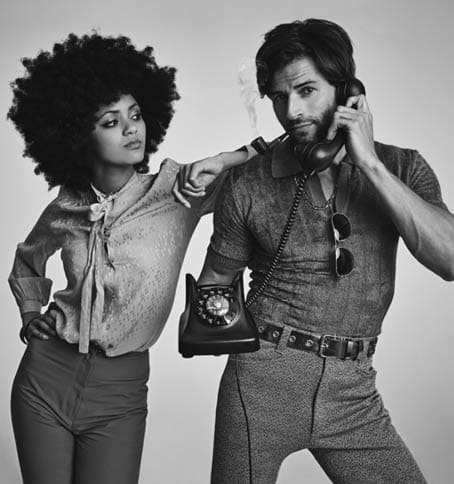 A beautiful young hippie looking over at a handsome man using a retro telephone.