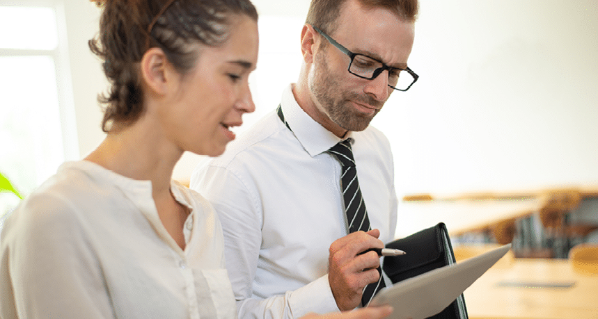 Business woman showing something on tablet screen to thoughtful male colleague. Woman and man standing in office with blurred interior in background. Business issue concept.