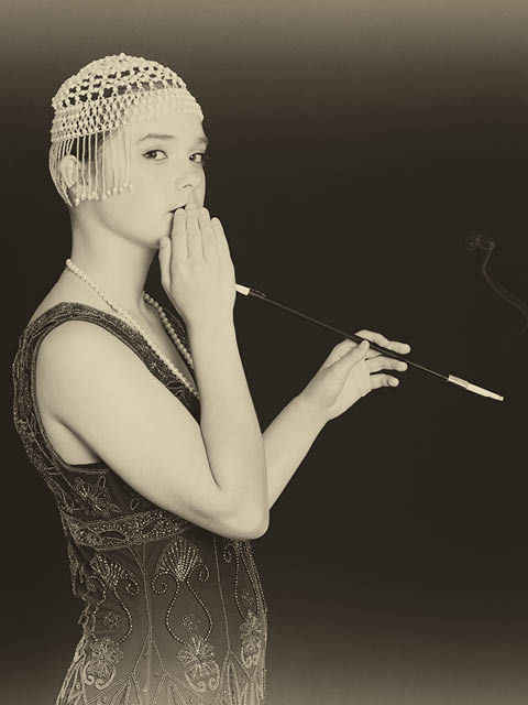 Black and white sepia toned photo of a teen flapper girl on a black background with a cigarette in a long cigarette filter
