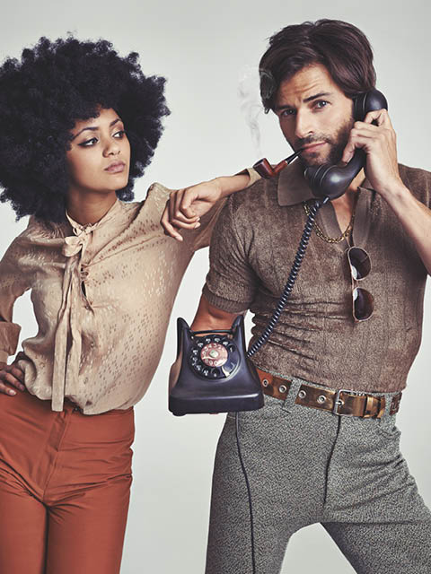 A beautiful young hippie looking over at a handsome man using a retro telephone.